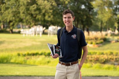 A student holding two books and a backpack outside by the patio. 